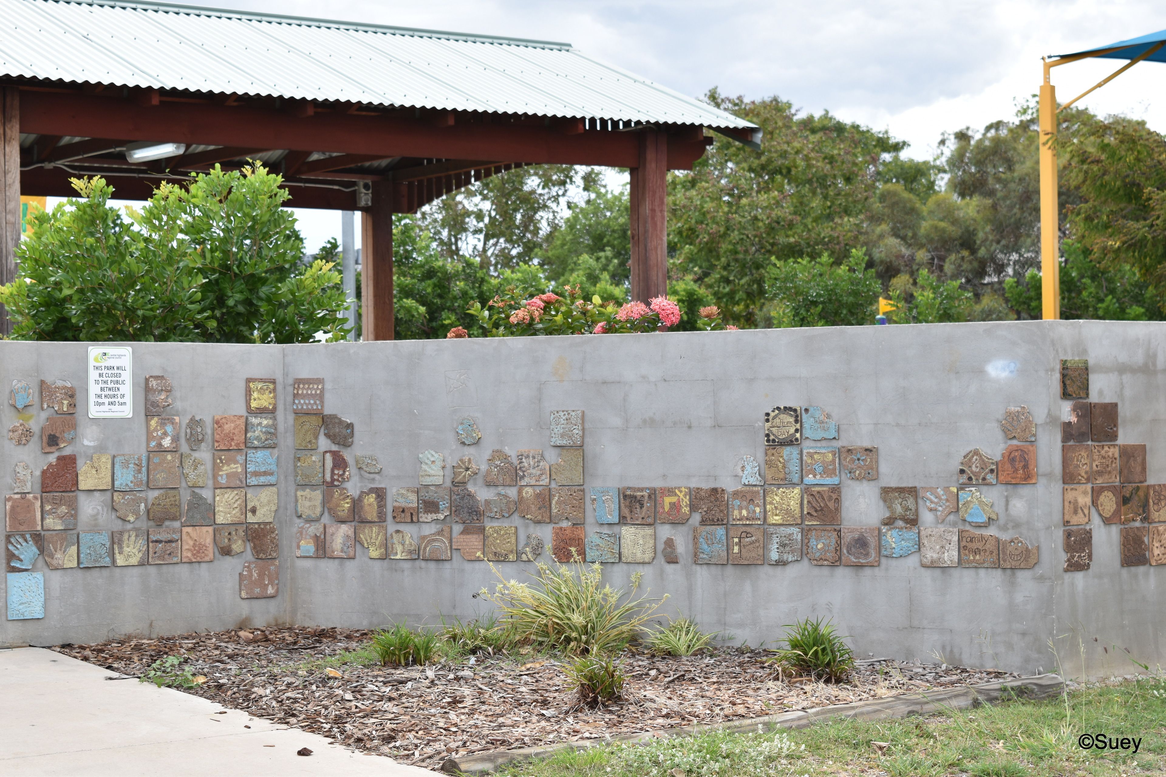 A concrete wall with a mosaic of tiles, a metal roofed structure, and a garden area with plants.