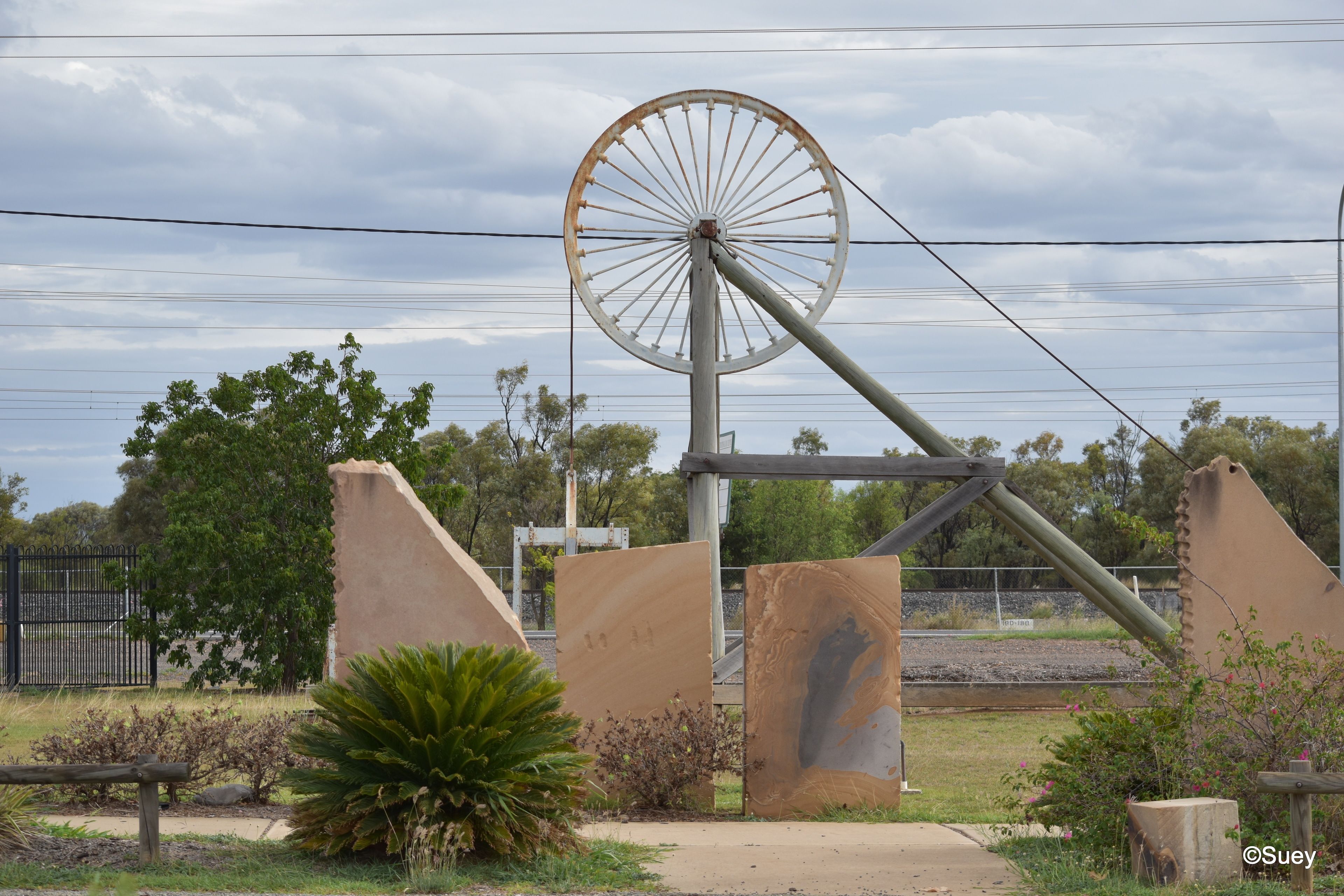 A large mechanical structure with a wheel and metal beams, surrounded by greenery and a fence.