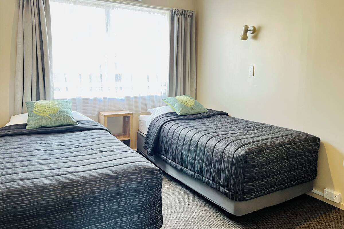 A hotel room with two beds, a window with curtains, an air conditioning unit, a bedside table, and a wall-mounted light switch.