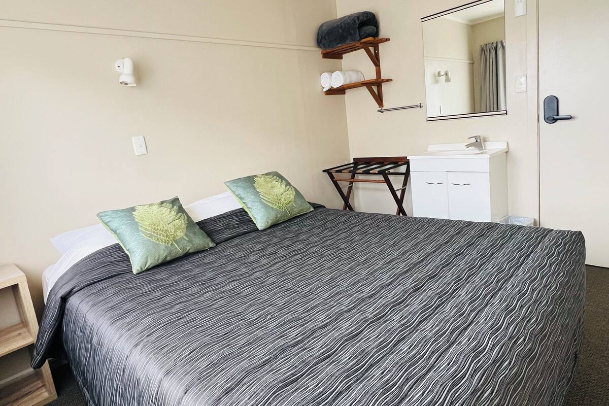 A bedroom with a bed, pillows, a small table, a mirror, a shelf with towels, and a white cabinet.