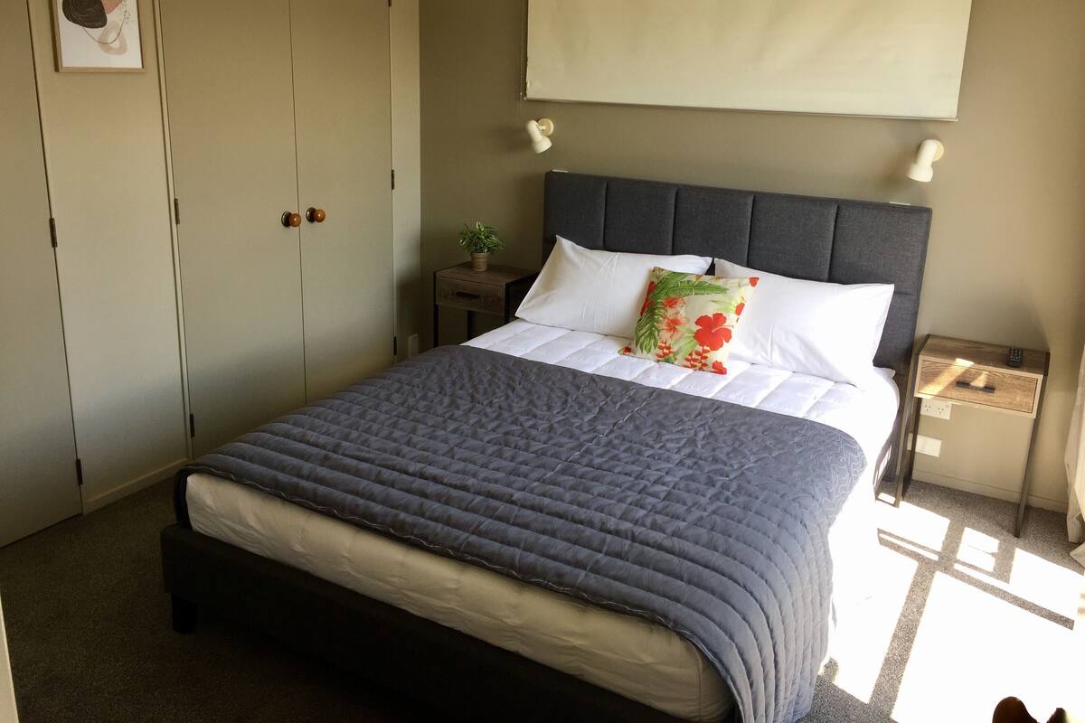 A neatly made bed with a grey quilt, a floral pillow, and a white pillow. There is a small bedside table with a plant and a remote control. A grey headboard and a white curtain are visible.