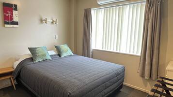 Cama bem arrumada com colcha cinza, duas almofadas verdes, mesa de cabeceira com abajur e janela com cortinas.