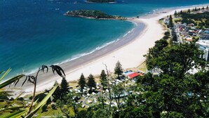 A coastal view with a beach, a small island, and a town with buildings and parked cars. - Seagulls Guesthouse (Tauranga)