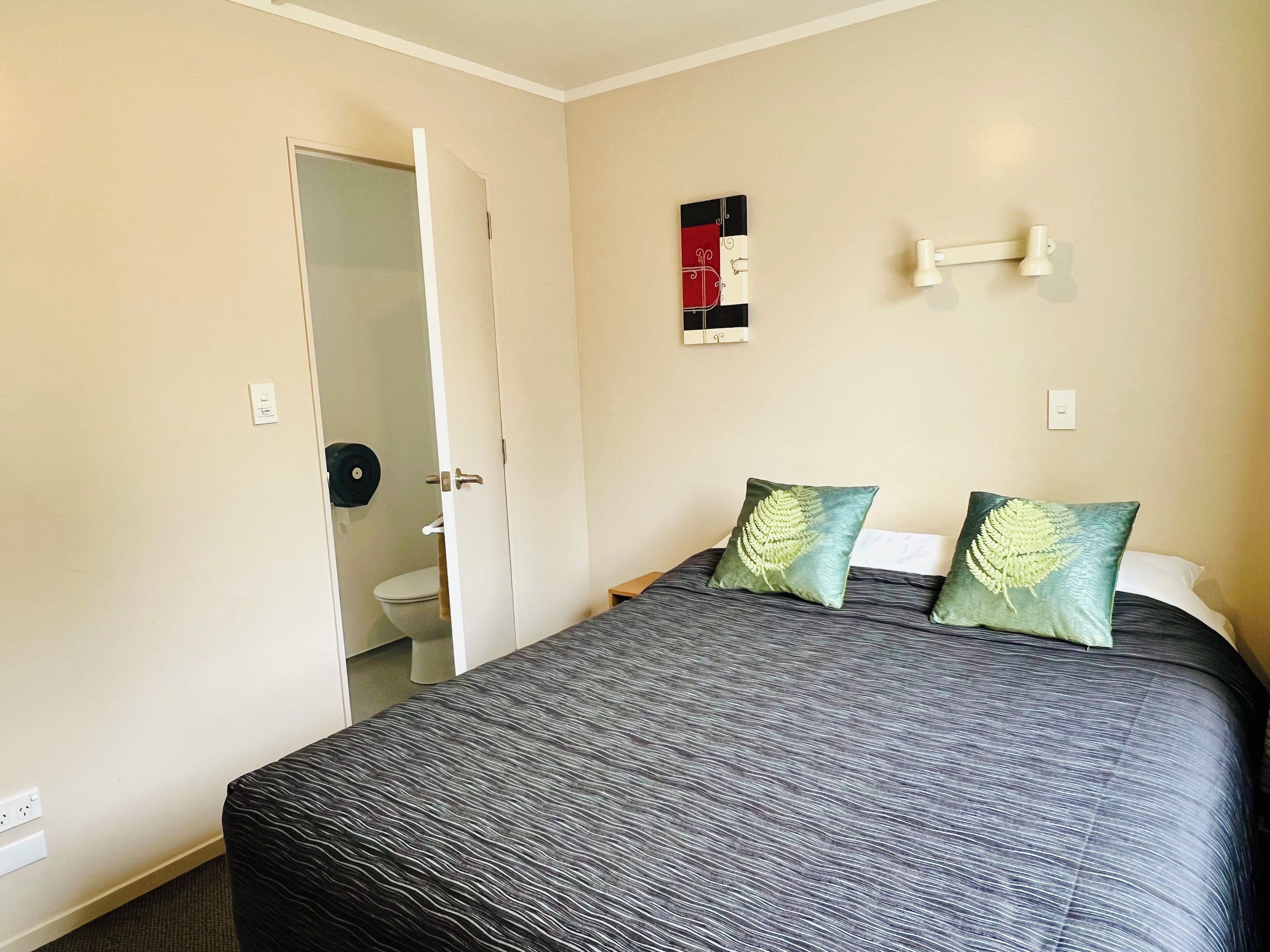A hotel room with a bed, two pillows, a wall-mounted light, a door leading to a bathroom, and a small artwork on the wall.