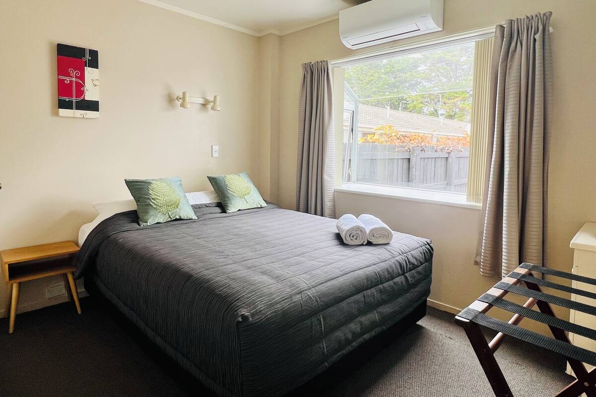 A hotel room with a bed, a bench, an air conditioner, and a window with curtains.