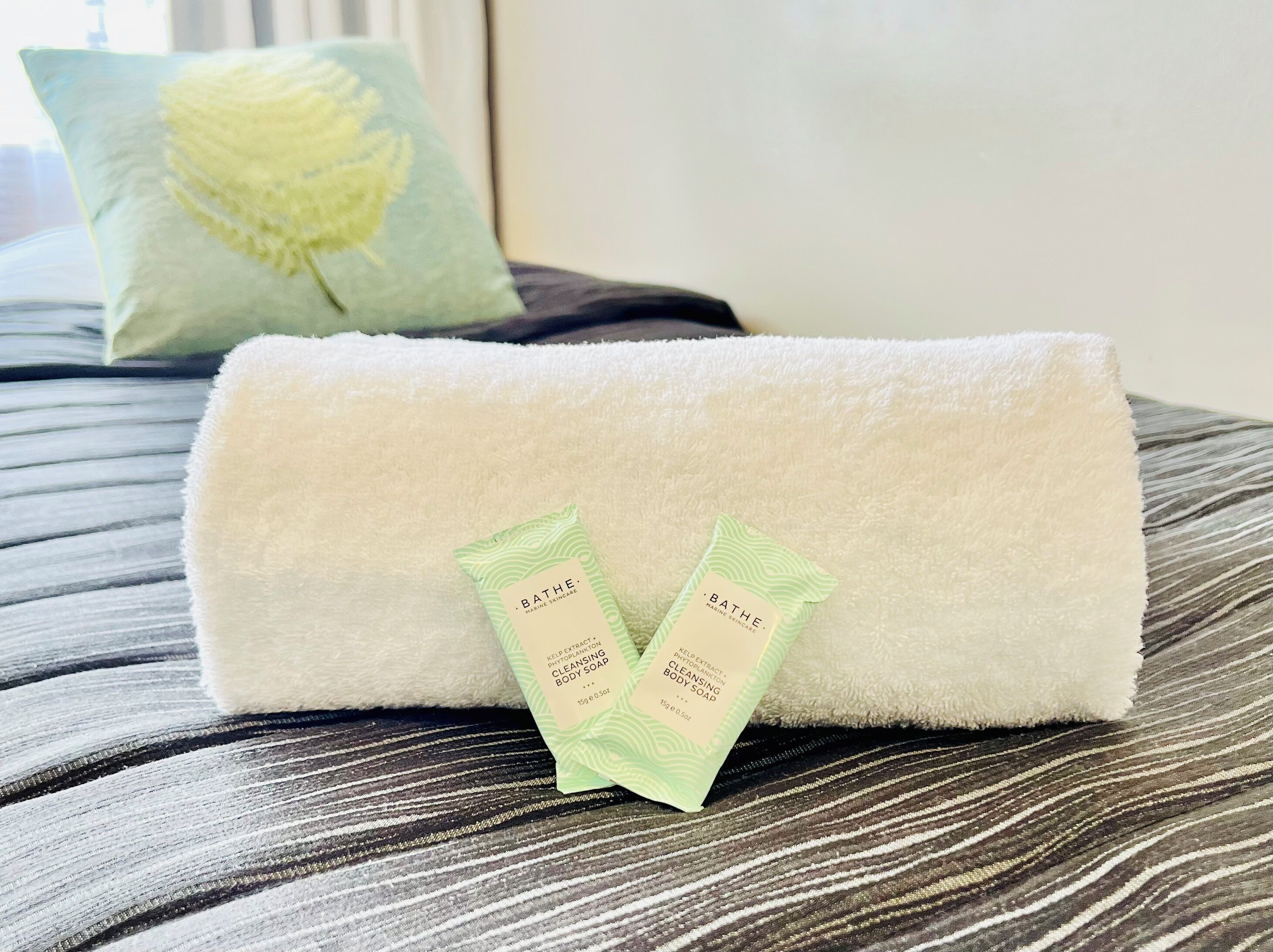 A neatly folded white towel on a bed with a striped pattern. Two green sachets of Bath & Body Works cleansing body soap are placed on the towel.