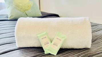 Una toalla blanca doblada con cuidado sobre una cama con estampado a rayas. Dos saqueles verdes de Bath & Body Works con jabón limpiador para el cuerpo están colocados sobre la toalla.