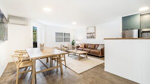 En moderne stue med et træspisebord, en brun lædersofa og et køkkenområde med hvide skabe samt et rustfrit stål køleskab.