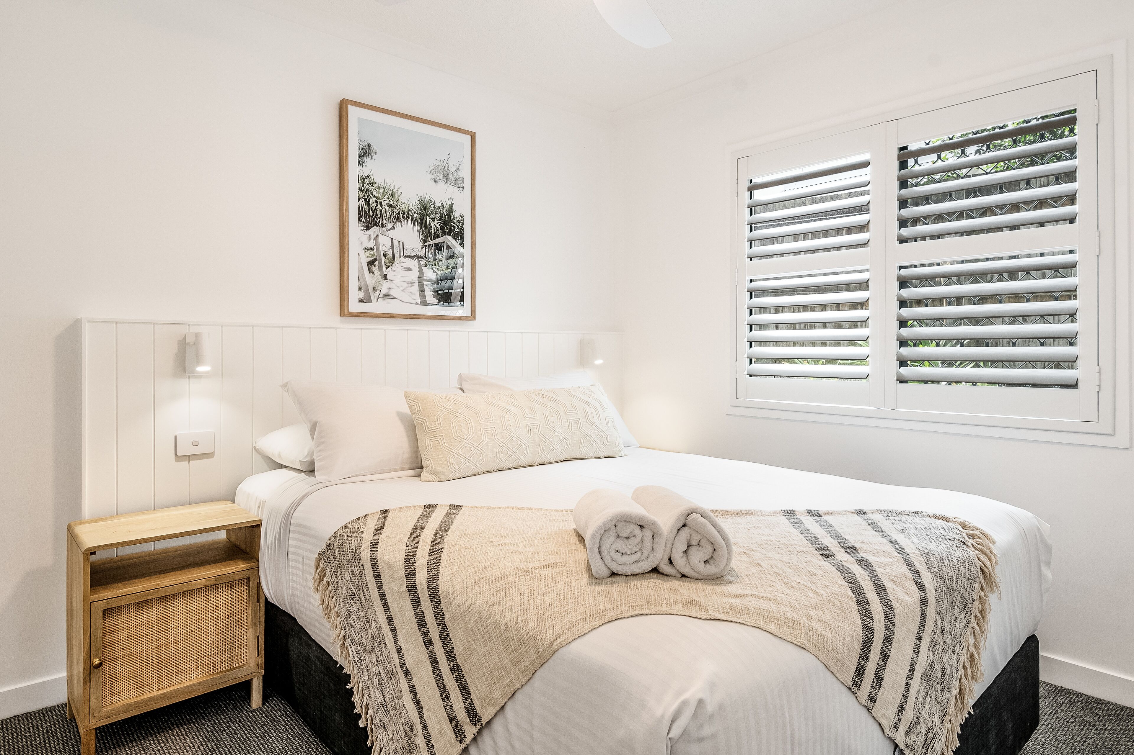 A neatly made bed with a striped blanket, a bedside table with a woven basket, and a window with blinds.