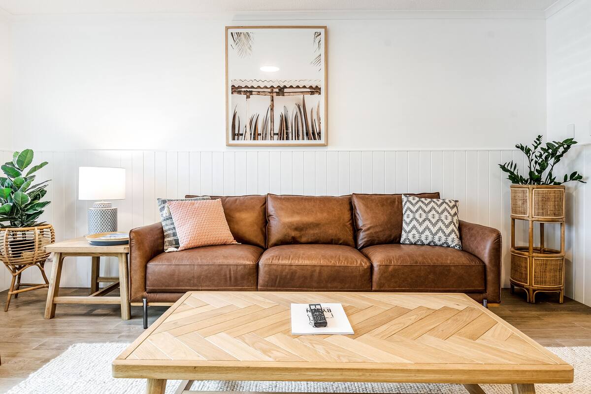 Un sofá de cuero marrón con cojines estampados, una mesa de centro de madera con un control remoto, una mesita auxiliar de mimbre y un marco de fotos colgado en la pared.