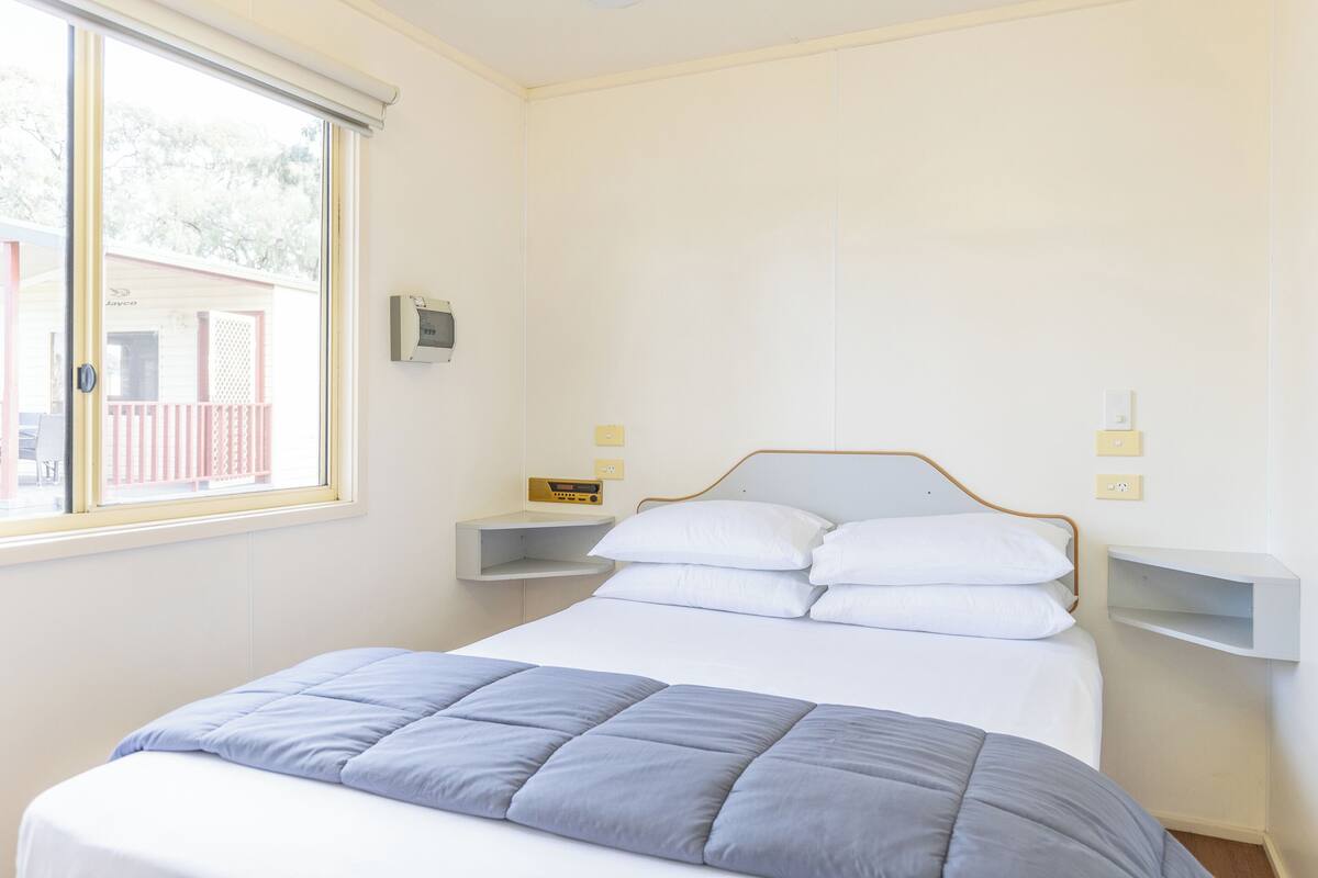 Uma cama bem arrumada com um edredom cinza e branco, uma mesa de cabeceira com abajur e uma janela com vista para uma cerca.