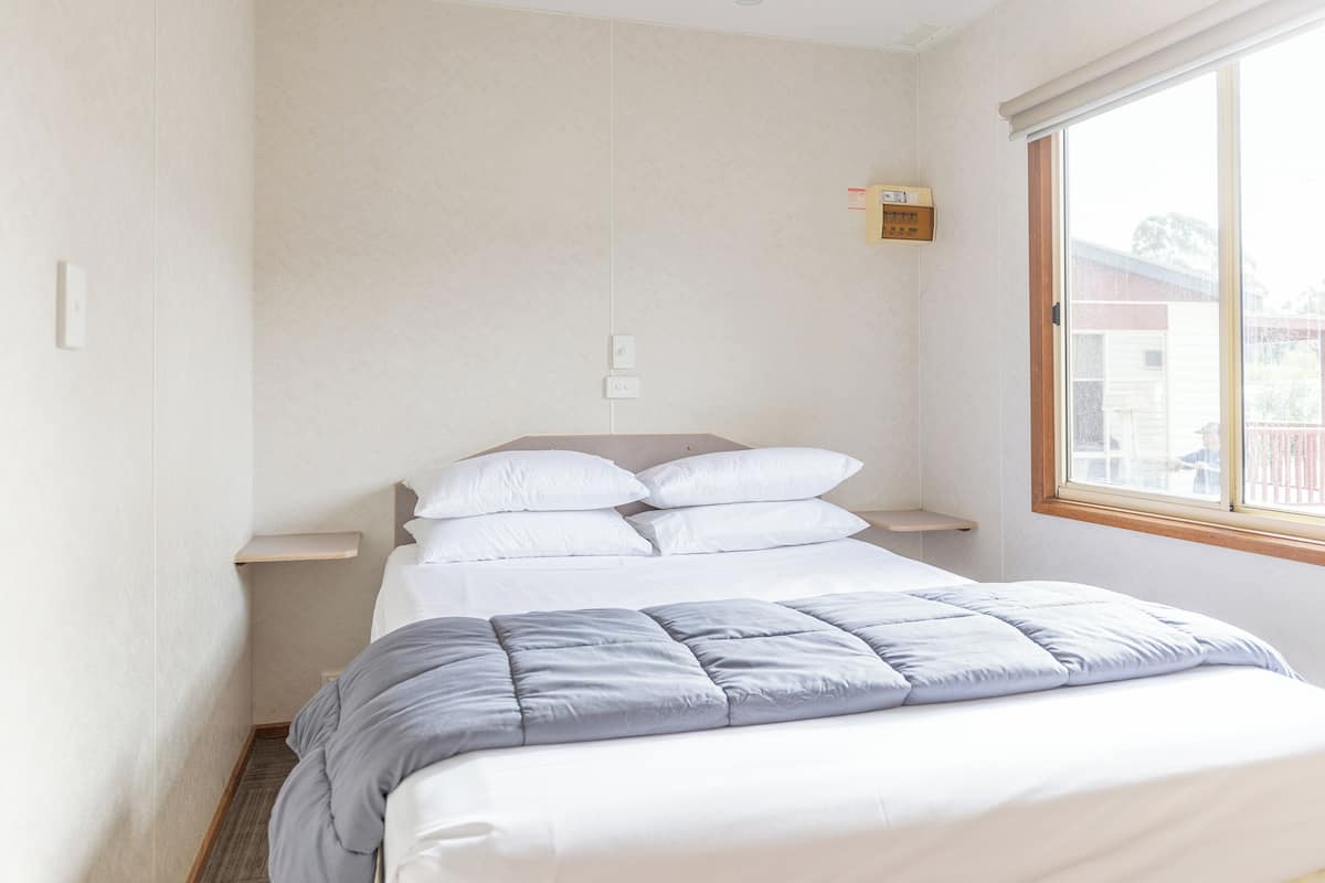 Cama bem arrumada com edredom cinza e almofadas brancas, mesa de cabeceira e janela com vista para uma casa.