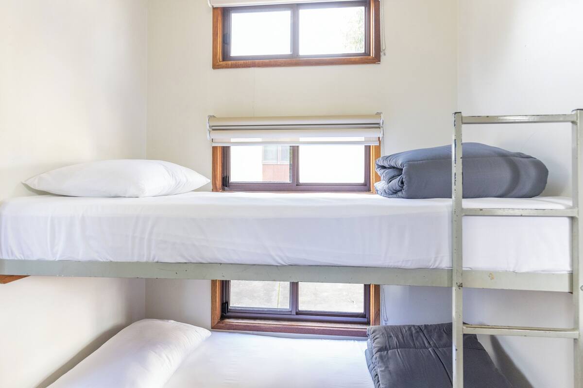Quarto com beliche, janela, roupa de cama branca e escada de metal.