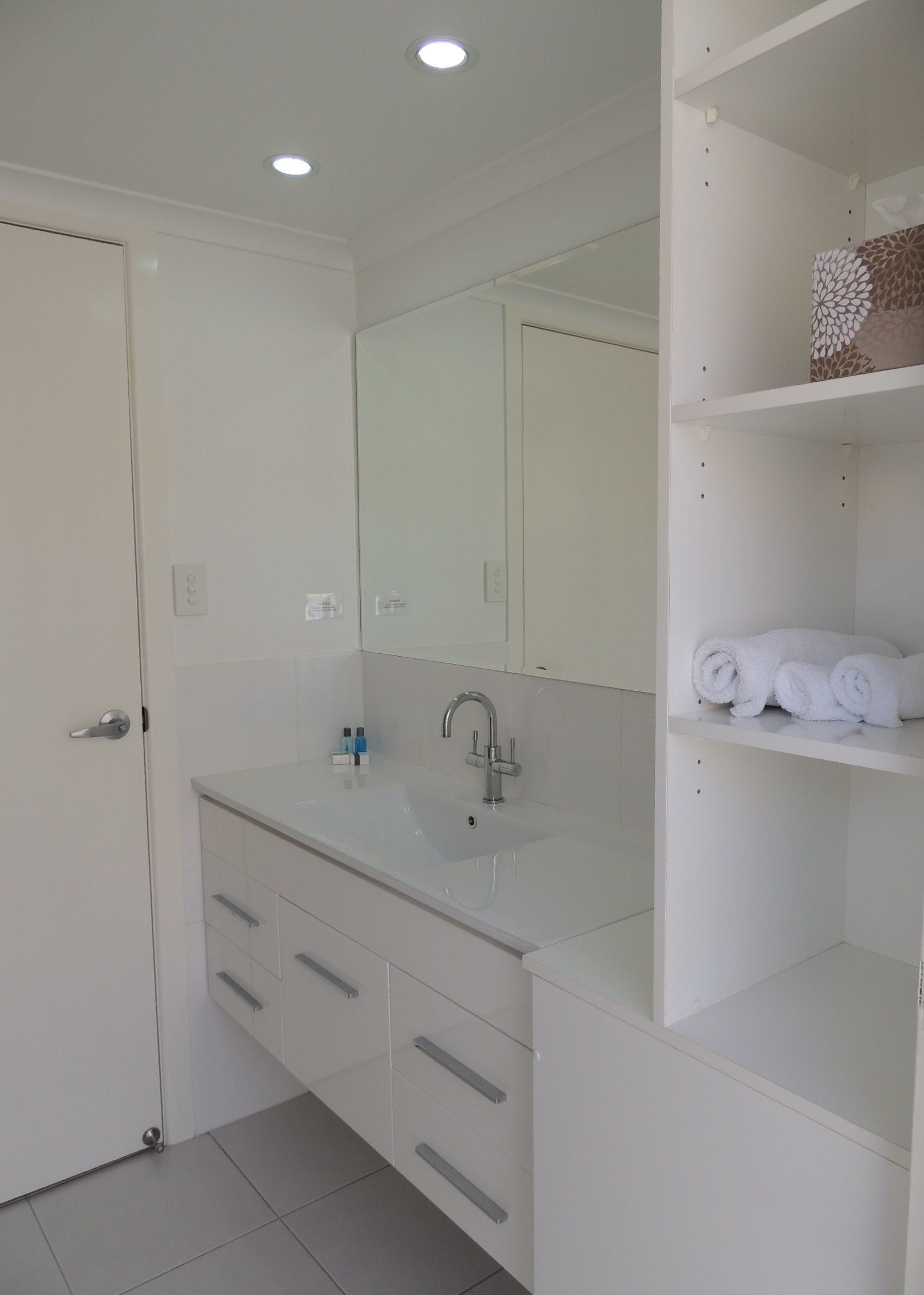Un baño moderno con un lavamanos blanco, espejo y estantes integrados.