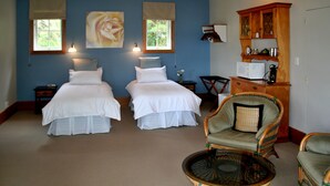 A room with two beds, a wicker chair, a small table, and a cabinet with shelves. - Villa Russell (Russell)