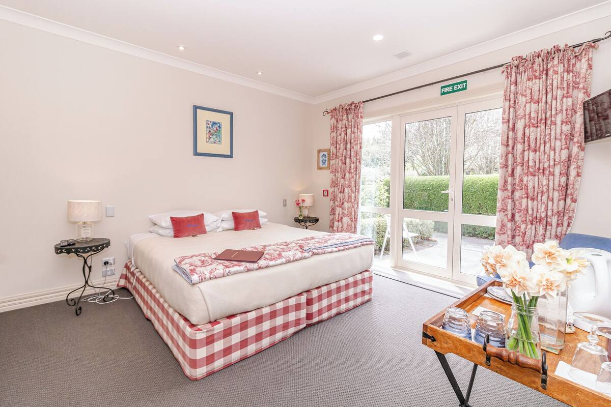 A hotel room with a bed, a bedside table with a lamp, a window with curtains, and a view of greenery outside.