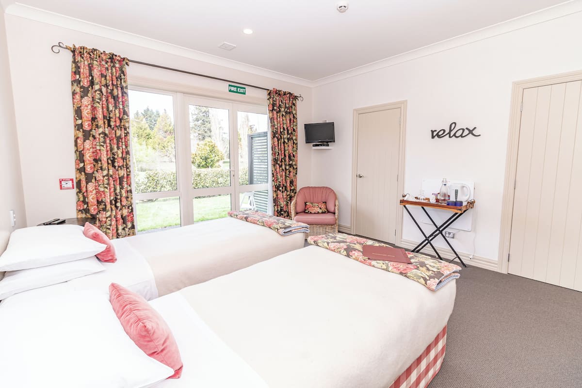 A hotel room with two beds, a chair, a small table with a kettle, and a sign indicating a fire exit.