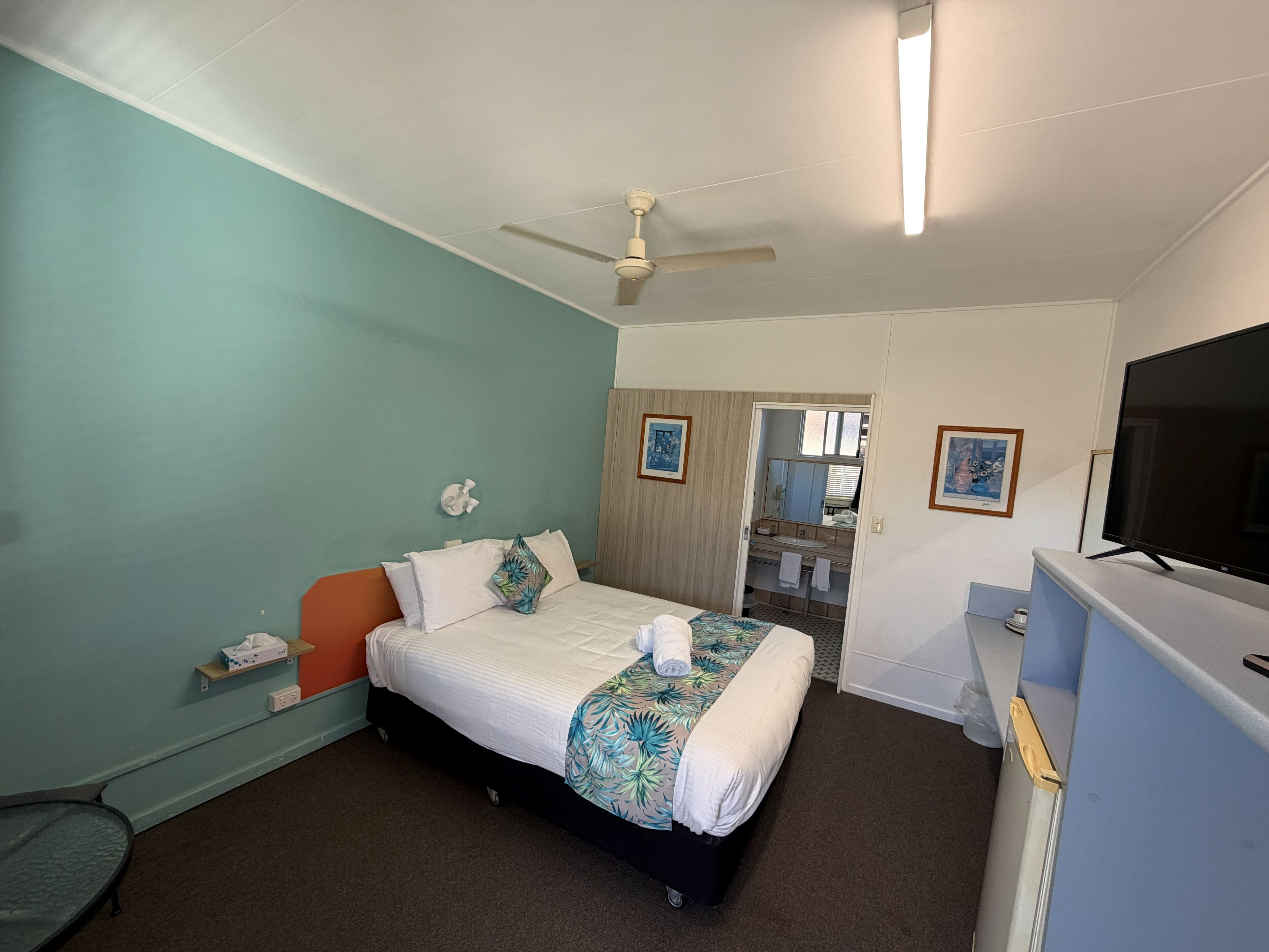 A hotel room with a bed, a ceiling fan, a TV, and a bathroom visible through an open door.