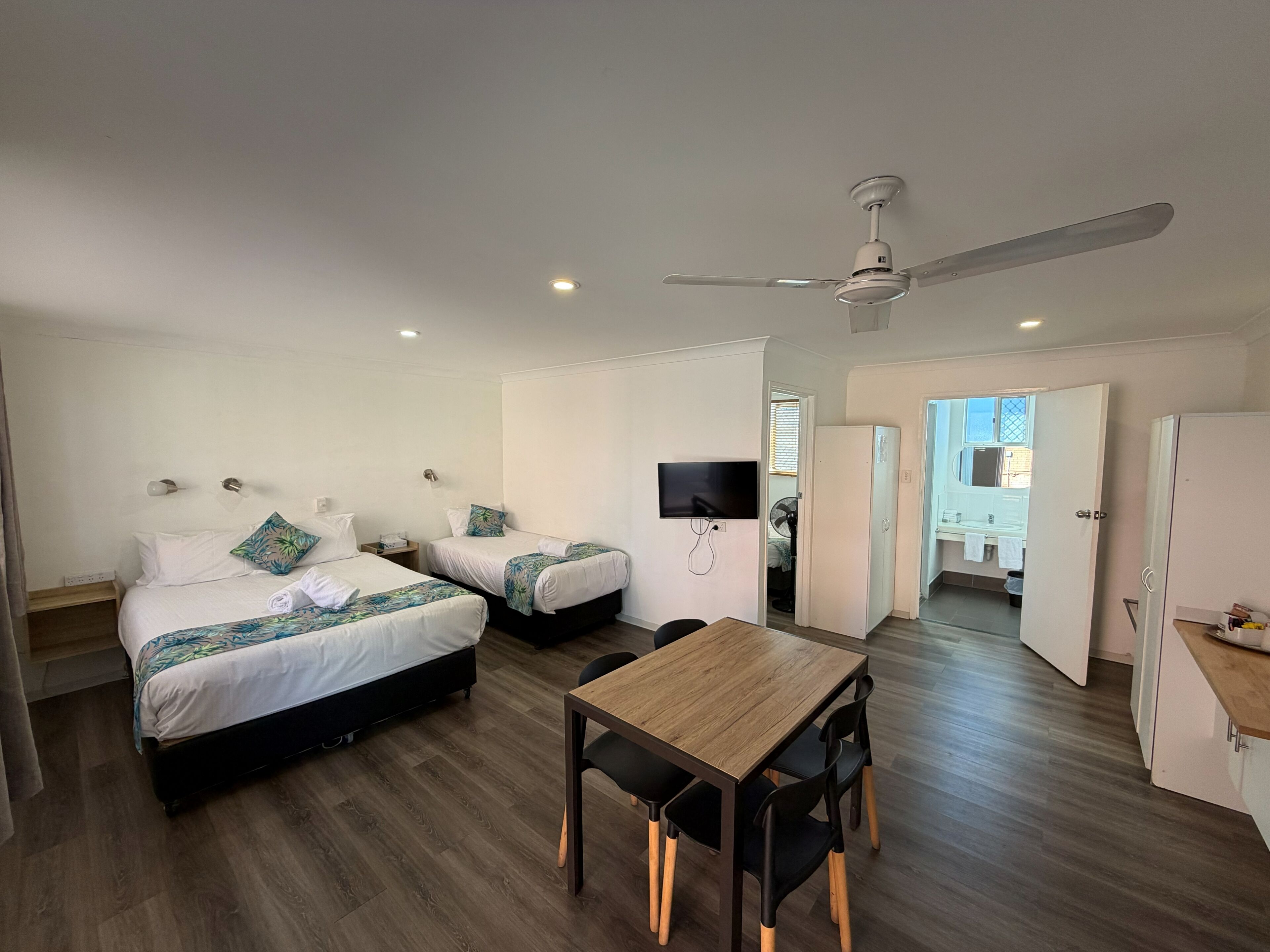 A hotel room with two beds, a dining table, a TV, and a ceiling fan.