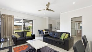 A modern living room with two black leather sofas, a white coffee table, and a glass dining table. There is a ceiling fan, a sliding glass door leading to a balcony, and a kitchen area visible through an open door.
