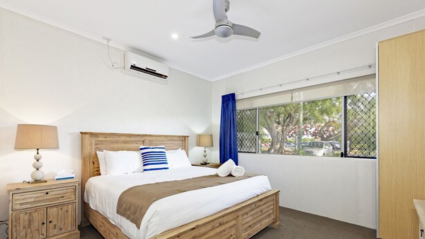 A bedroom with a wooden bed, a ceiling fan, an air conditioning unit, a nightstand with a lamp, and a window with a view of trees and parked cars.