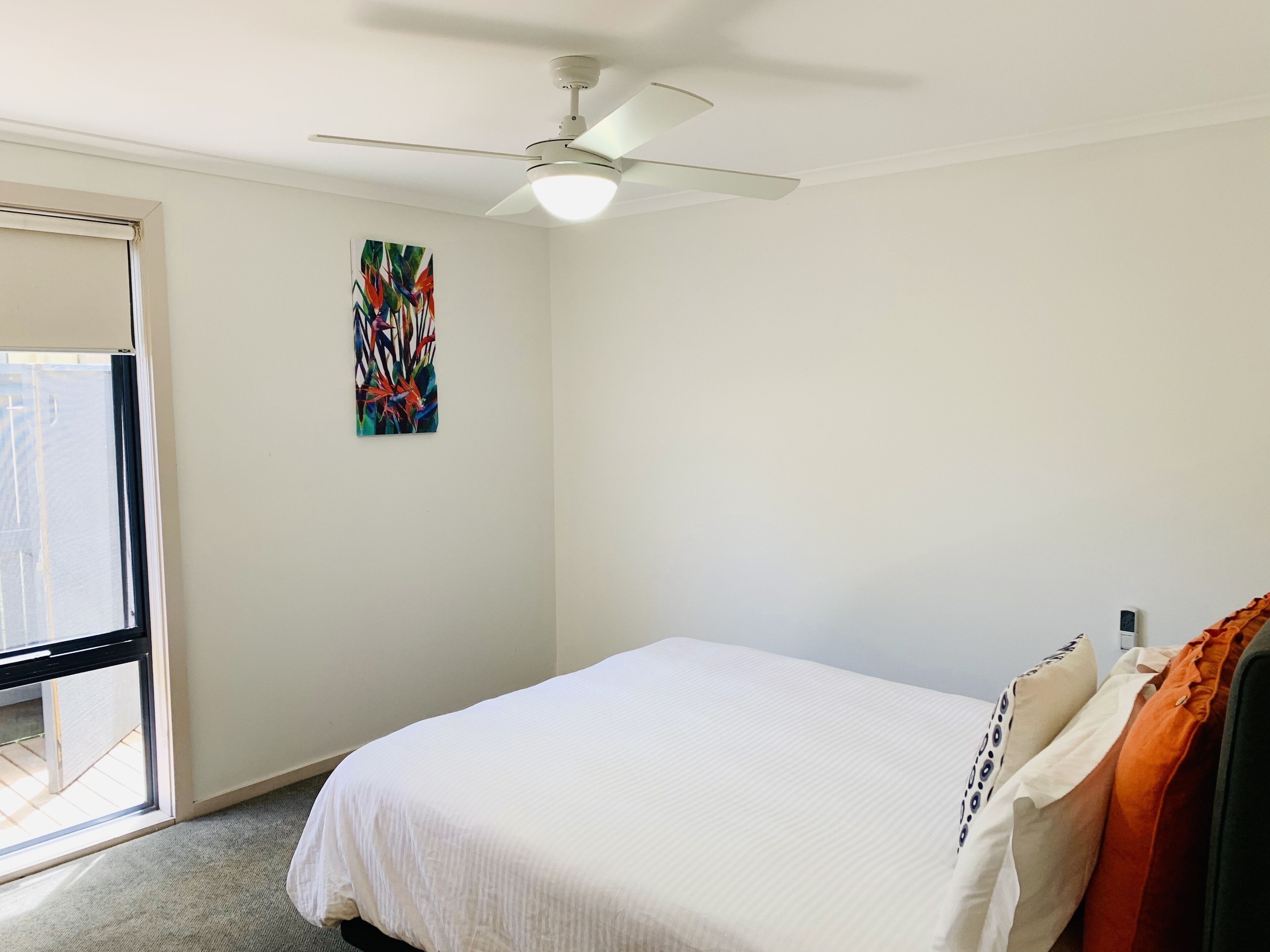 A bedroom with a bed, a ceiling fan, a window, and a wall-mounted artwork.