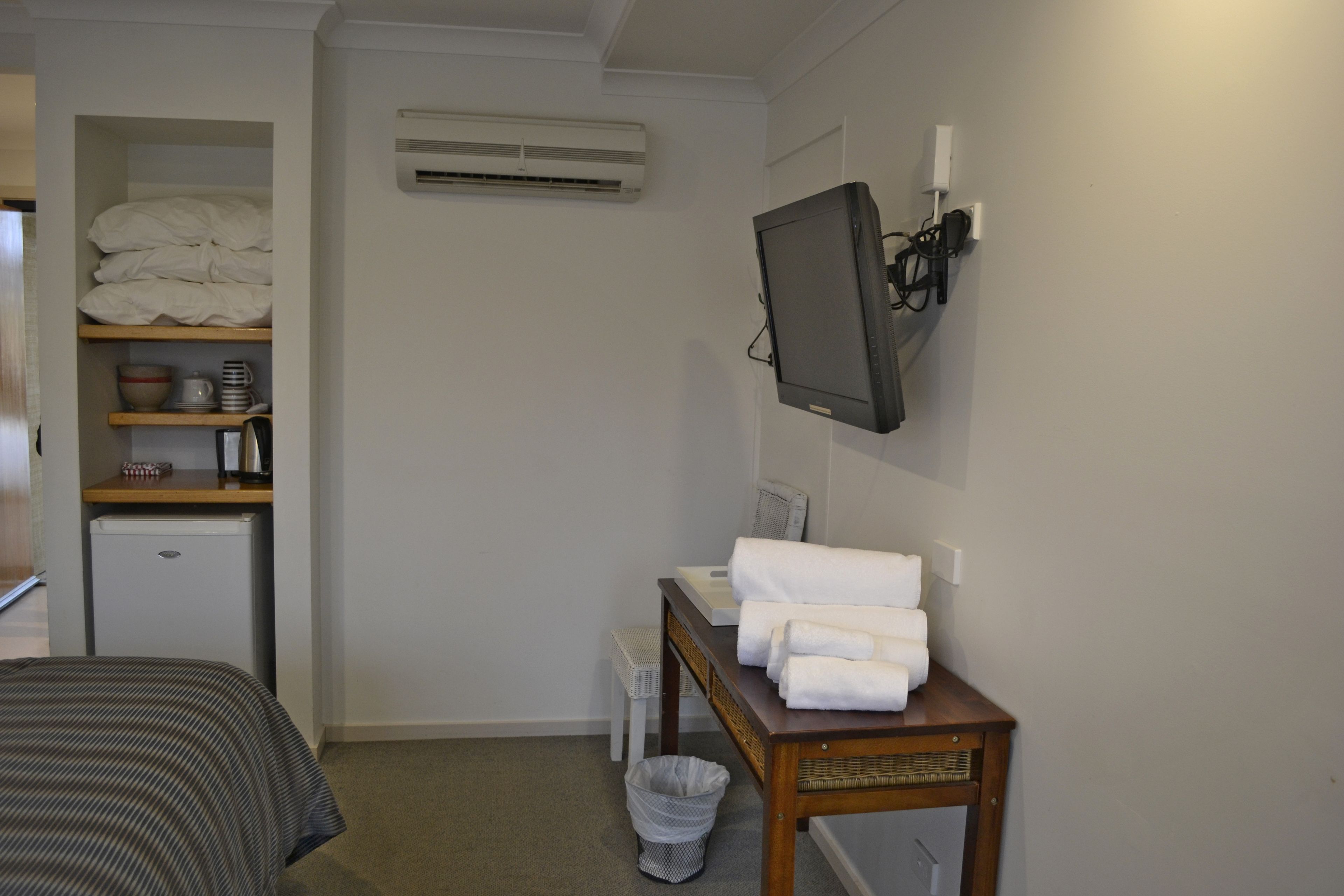 Una habitación de hotel pequeña con cama, escritorio con televisor, refrigerador y estante con artículos de aseo, además de un cesto de basura.