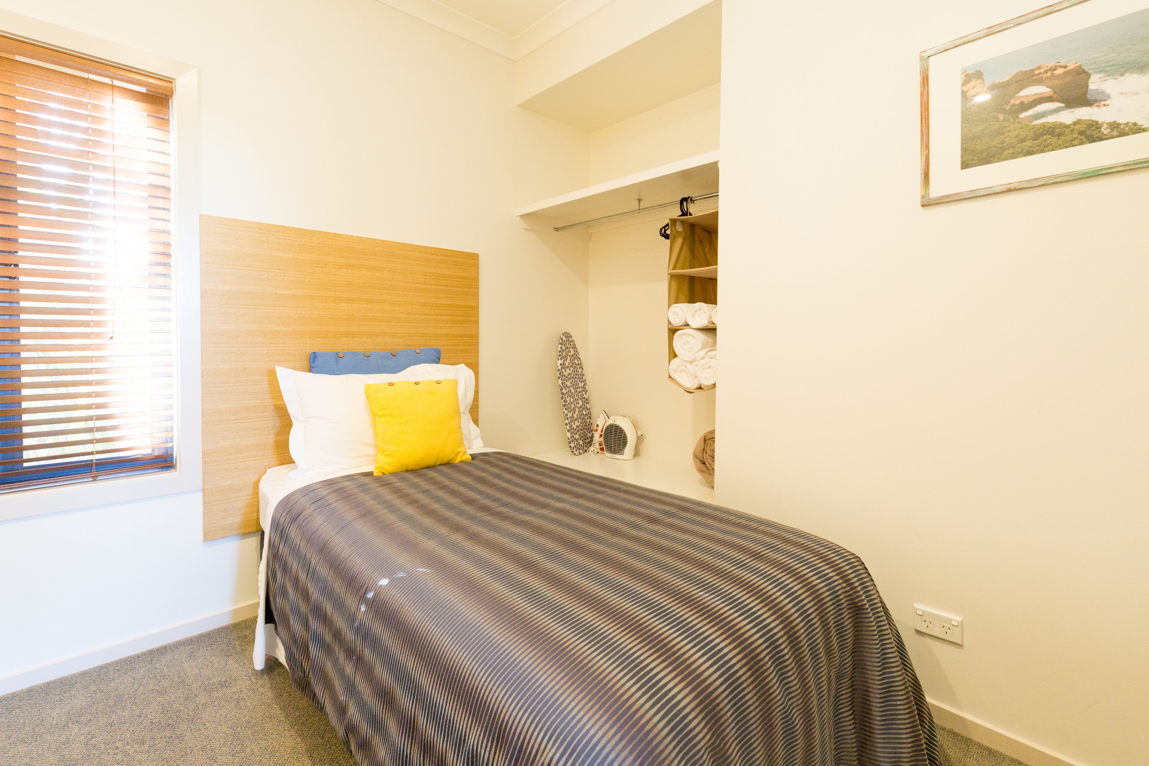 A bedroom with a bed, a window with blinds, a wall-mounted shelf with towels, and a framed picture.