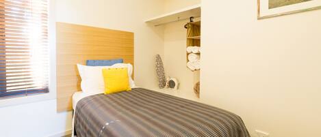 Un dormitorio con cama, ventana con persianas, estante en la pared con toallas e imagen enmarco.