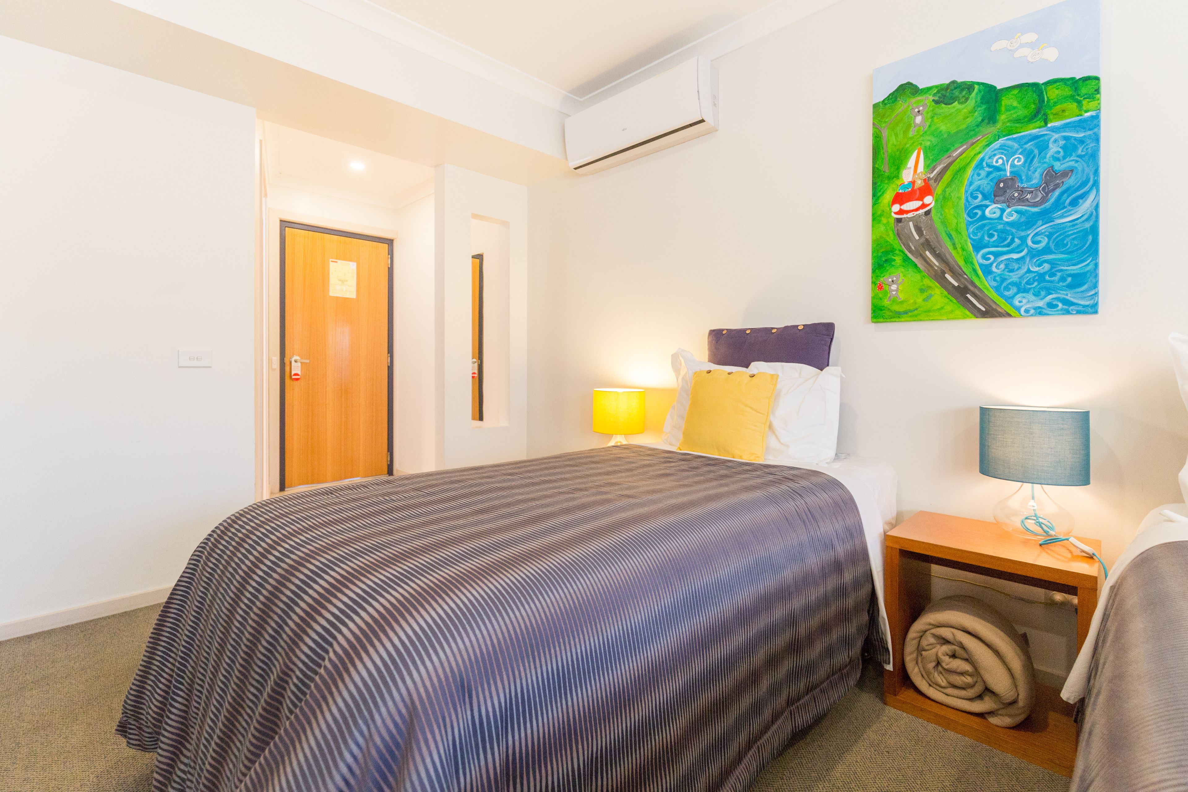 A hotel room with a bed, a nightstand with a lamp, a painting on the wall, and a door leading to another room.