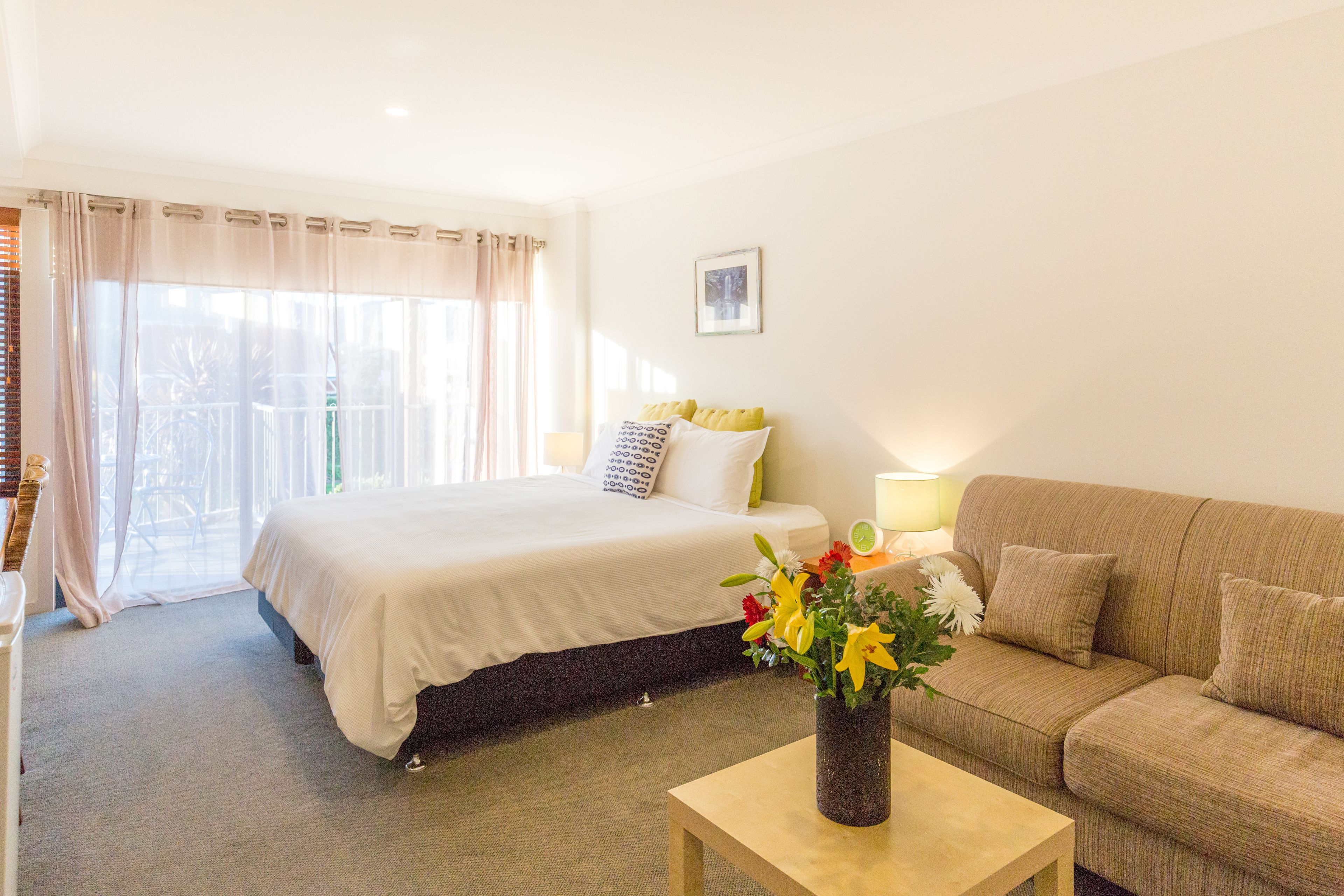 Una habitación de hotel con cama, sofá, una mesita de centro con un florero y una lámpara, además de un ventanal con cortinas.