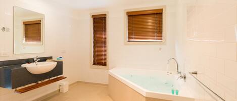 A modern bathroom with a large bathtub, a sink, and two windows with blinds.