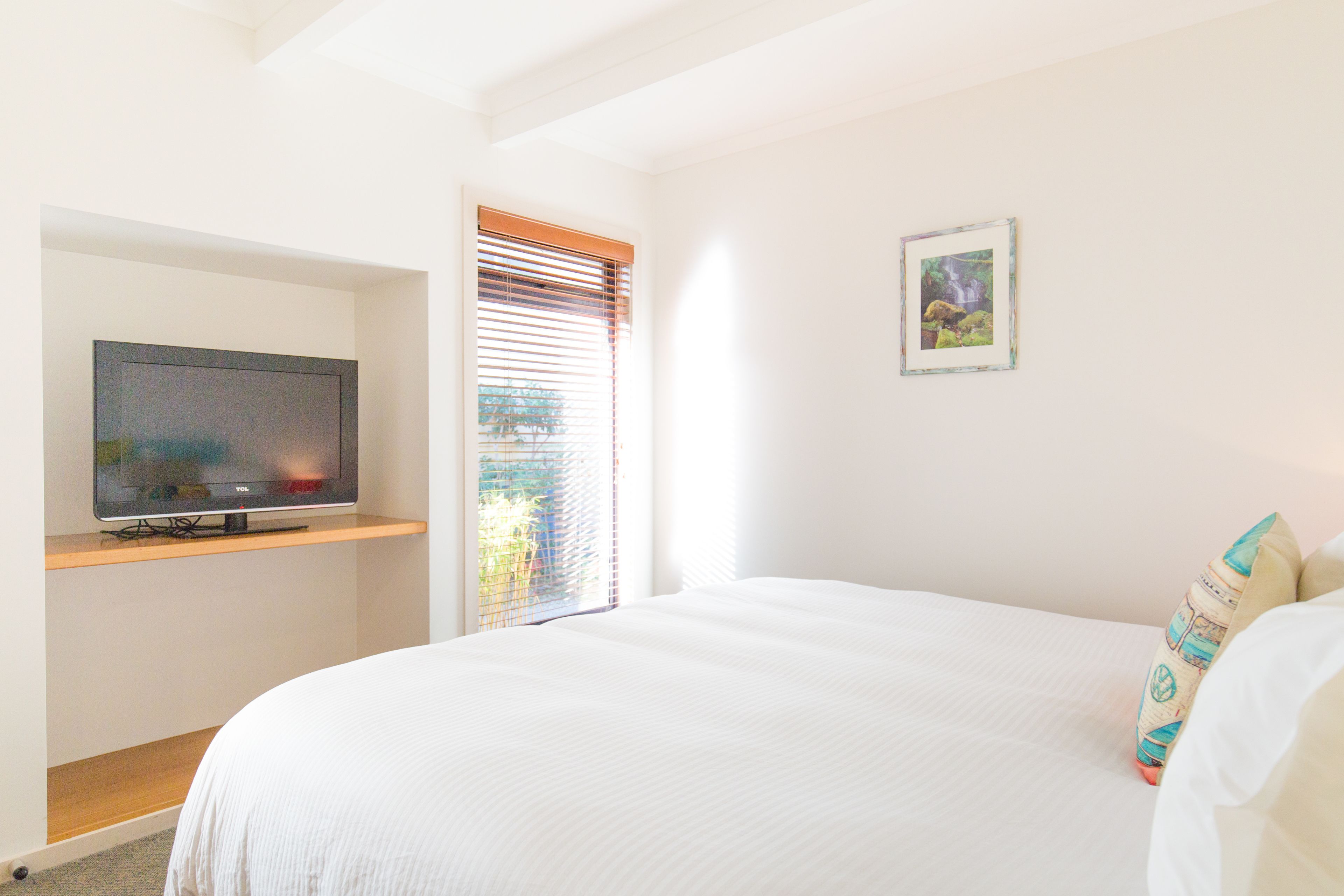 Un dormitorio con una cama, un televisor en un estante, una ventana con persianas y un cuadro en la pared.