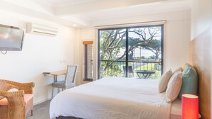 Une chambre d’hôtel avec un grand lit, un bureau avec une chaise, une télévision et un balcon donnant sur des arbres, avec une table.