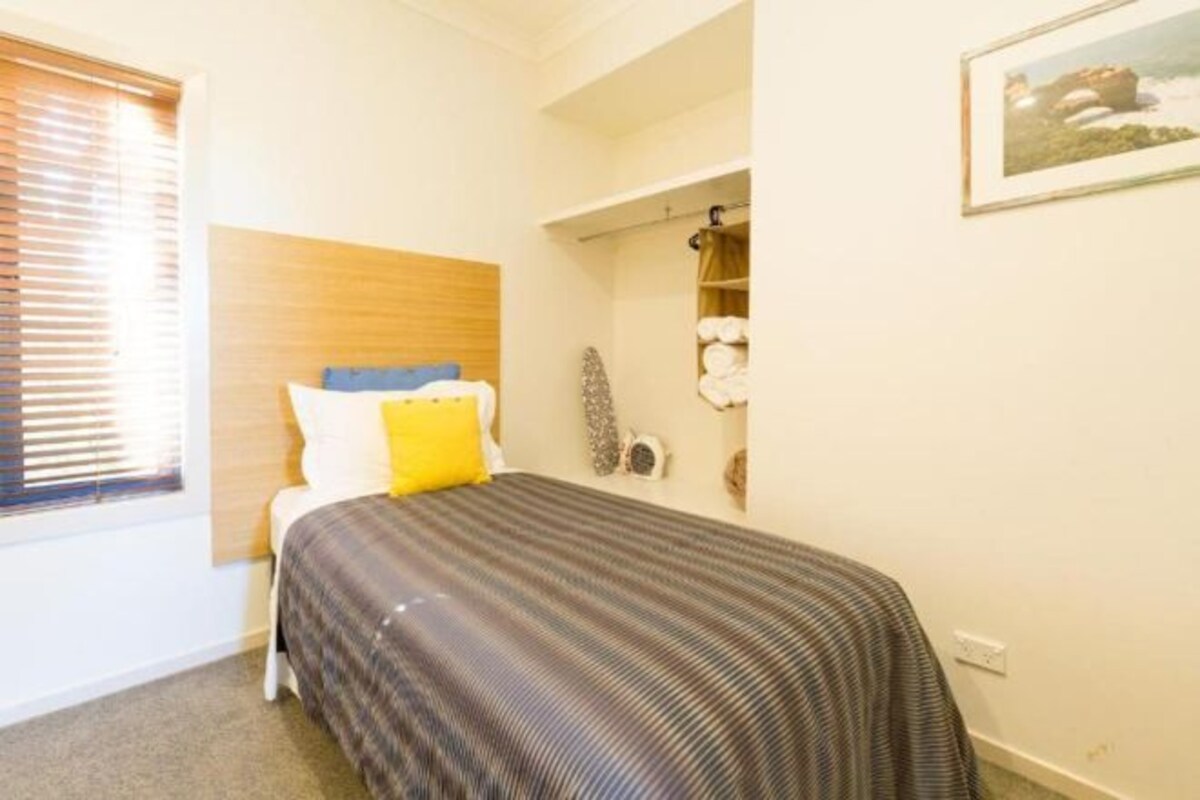 A bedroom with a bed, a window with blinds, a wall-mounted shelf with towels, and a framed picture.
