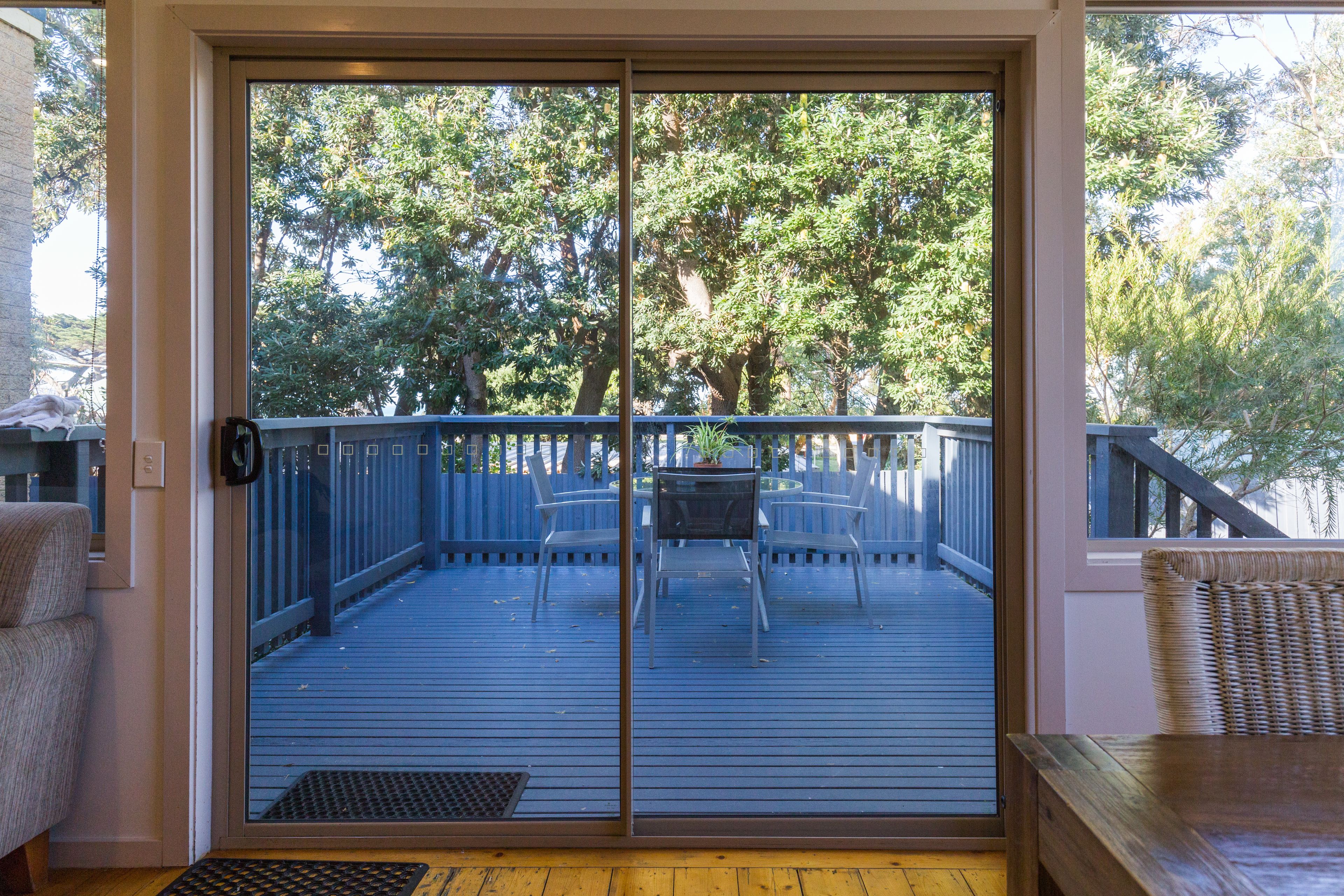 Una puerta corrediza de vidrio da a una terraza exterior con una mesa y sillas, rodeada por un piso de madera y una barandilla azul.