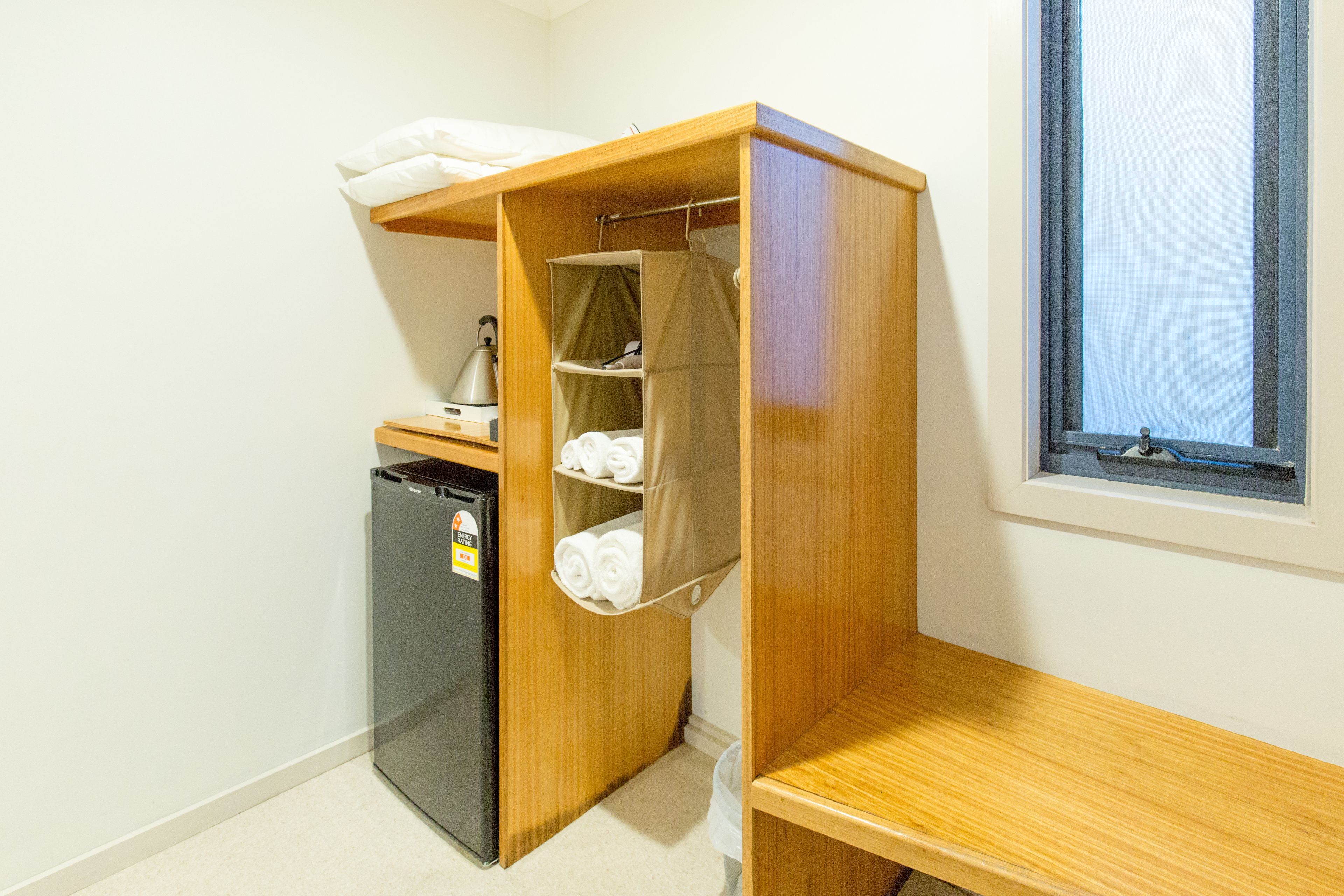 A compact room with a kitchenette, a small refrigerator, a wooden shelf unit with storage, and a window.