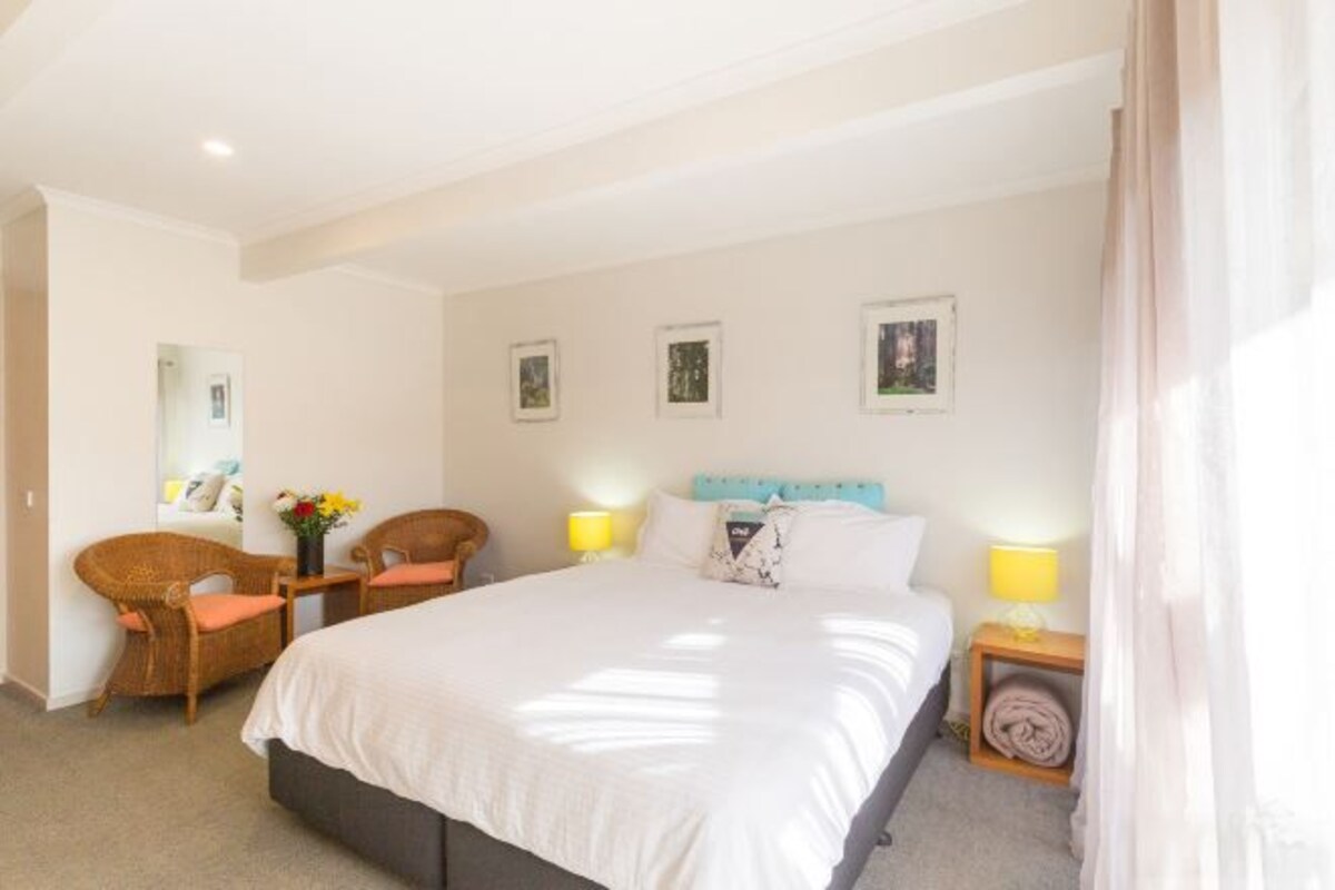 A hotel room with a large bed, two chairs, a small table with a vase of flowers, and a nightstand with a lamp.