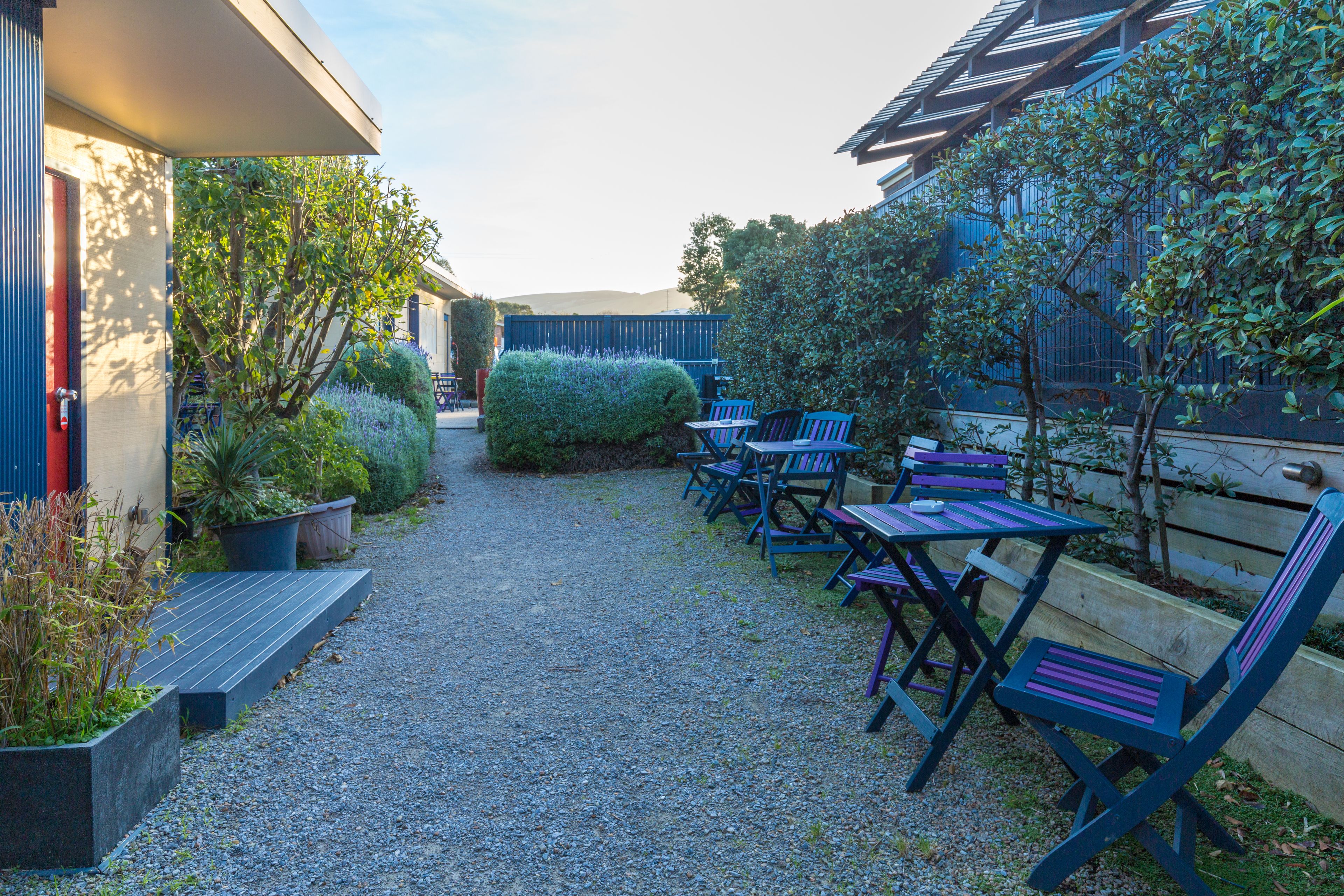 Un chemin de gravier mène à un espace extérieur aménagé avec des bancs et des chaises, entouré de verdure et bordé d’un bâtiment doté d’un toit en pente.