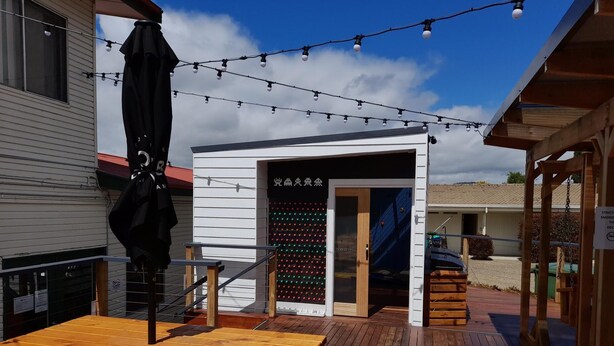 Una terraza de madera con mesas de picnic, un edificio pequeño con un letrero y luces colgantes.