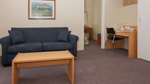 A living room with a sofa, a wooden coffee table, a desk with a chair, and a framed picture on the wall. - Cooma Motor Inn (Cooma)