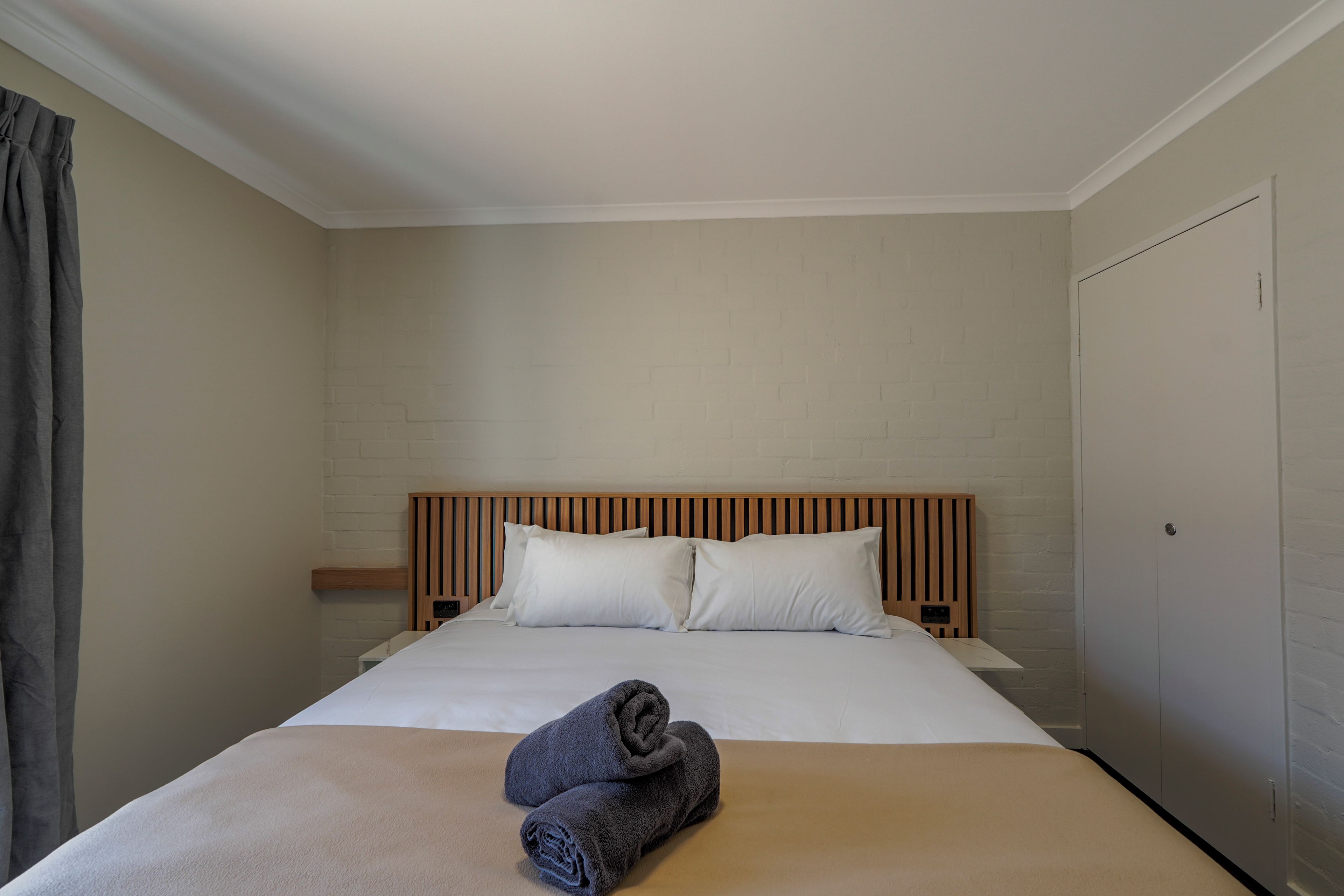 A hotel room with a large bed, two pillows, a wooden headboard, a nightstand, and a folded towel on the bed.