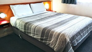 Una cama bien tendida con una colcha a rayas, una mesita de noche con lámpara y un enchufe en la pared.