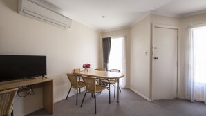 Habitaci ón de hotel con televisión de pantalla plana sobre un escritorio de madera, una mesa de comedor con cuatro sillas, una ventana con cortinas y una puerta.