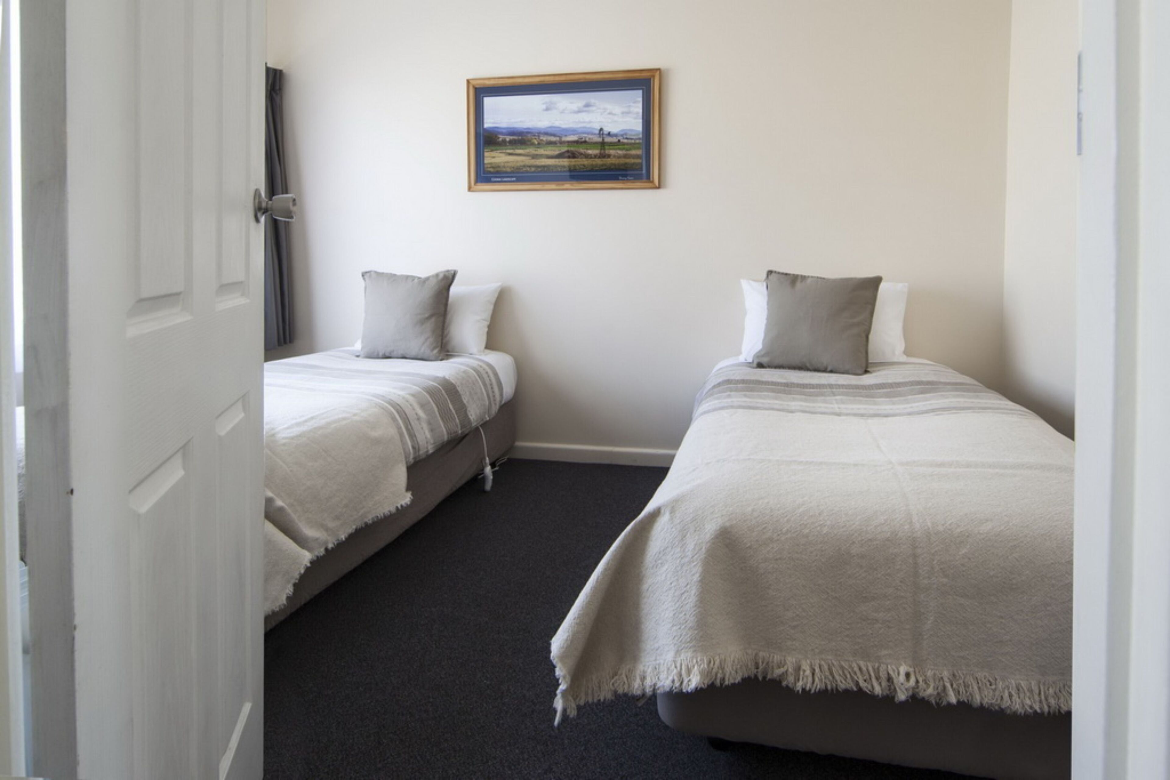 A room with two single beds, a framed landscape painting on the wall, and a door leading to another room.