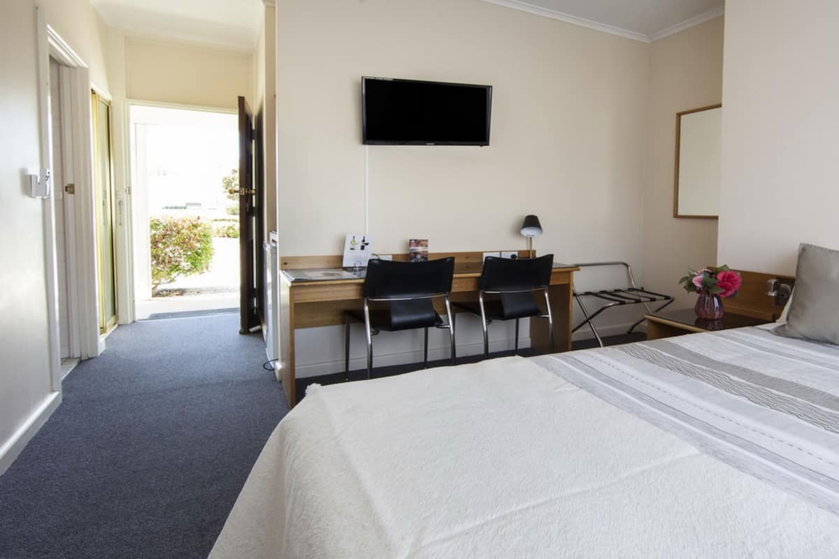 A hotel room with a bed, a desk with two chairs, a TV mounted on the wall, and a window with a view of greenery.