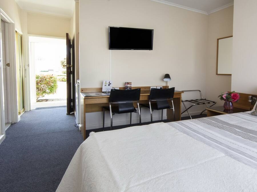 A hotel room with a bed, a desk with two chairs, a TV mounted on the wall, and a window with a view of greenery.