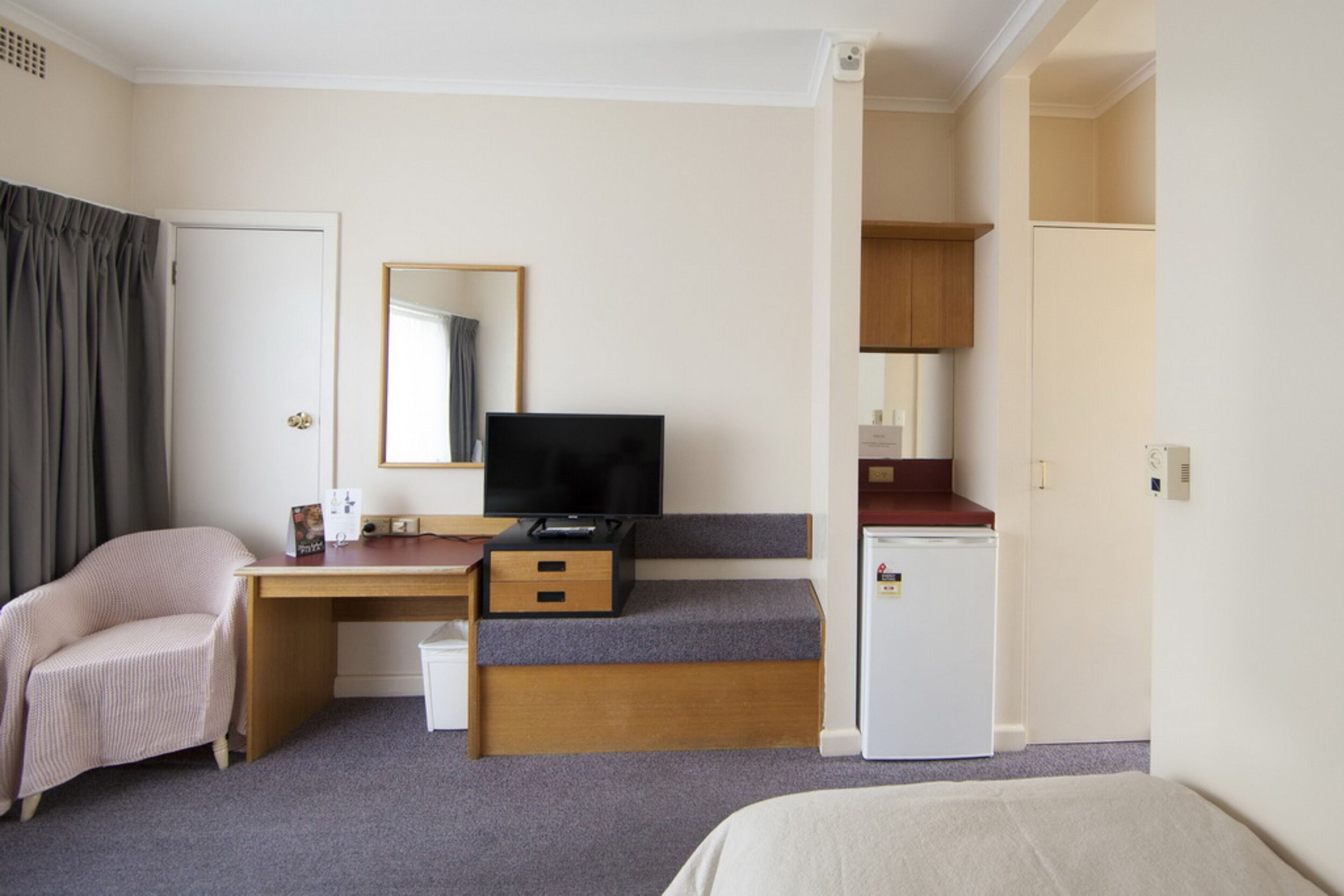 A hotel room with a bed, a desk with a television, a small refrigerator, and a kitchenette area.