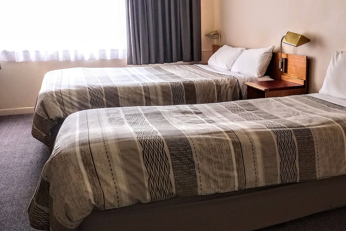 A hotel room with two beds, each with a patterned bedspread, a bedside table with a lamp, and a window with curtains.