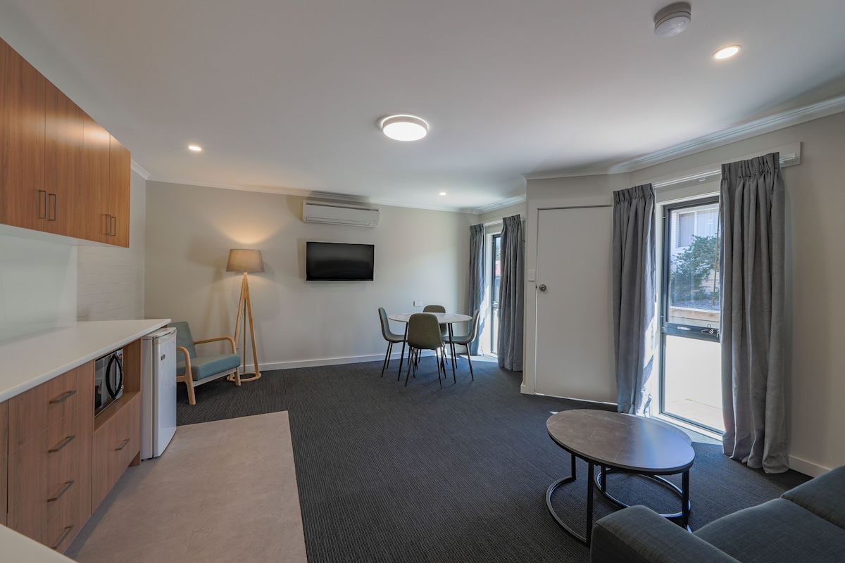 A modern hotel room with a kitchenette, a dining area, a sofa, and a large window with curtains.
